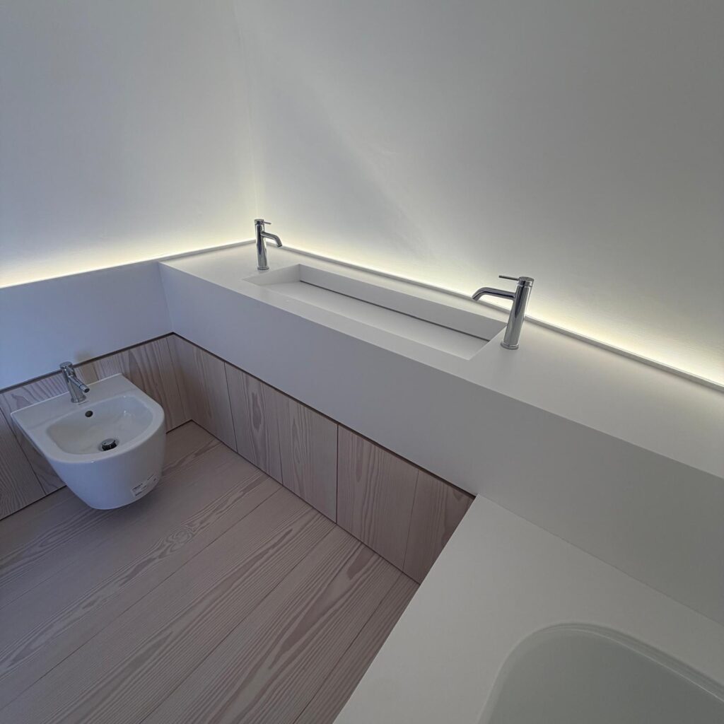 White Corian washbasin trough set into a continuous worktop, with two chrome pillar taps and timber-fronted cabinets