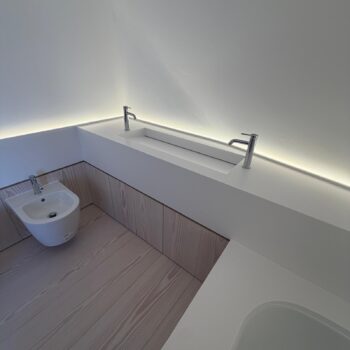White Corian washbasin trough set into a continuous worktop, with two chrome pillar taps and timber-fronted cabinets